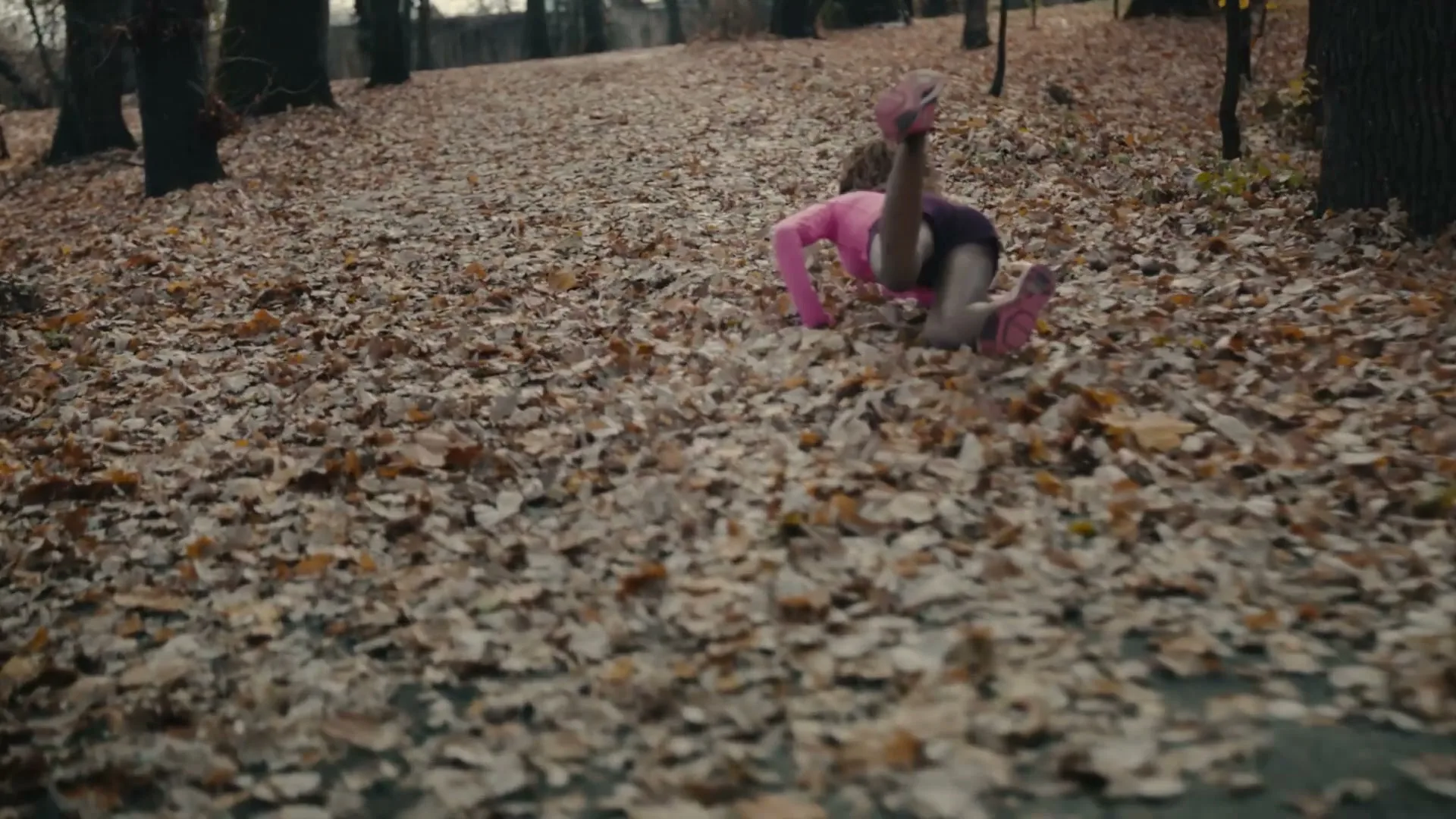 A woman in a pink top and dark shorts runs on a path covered in autumn leaves, then trips and falls forward.