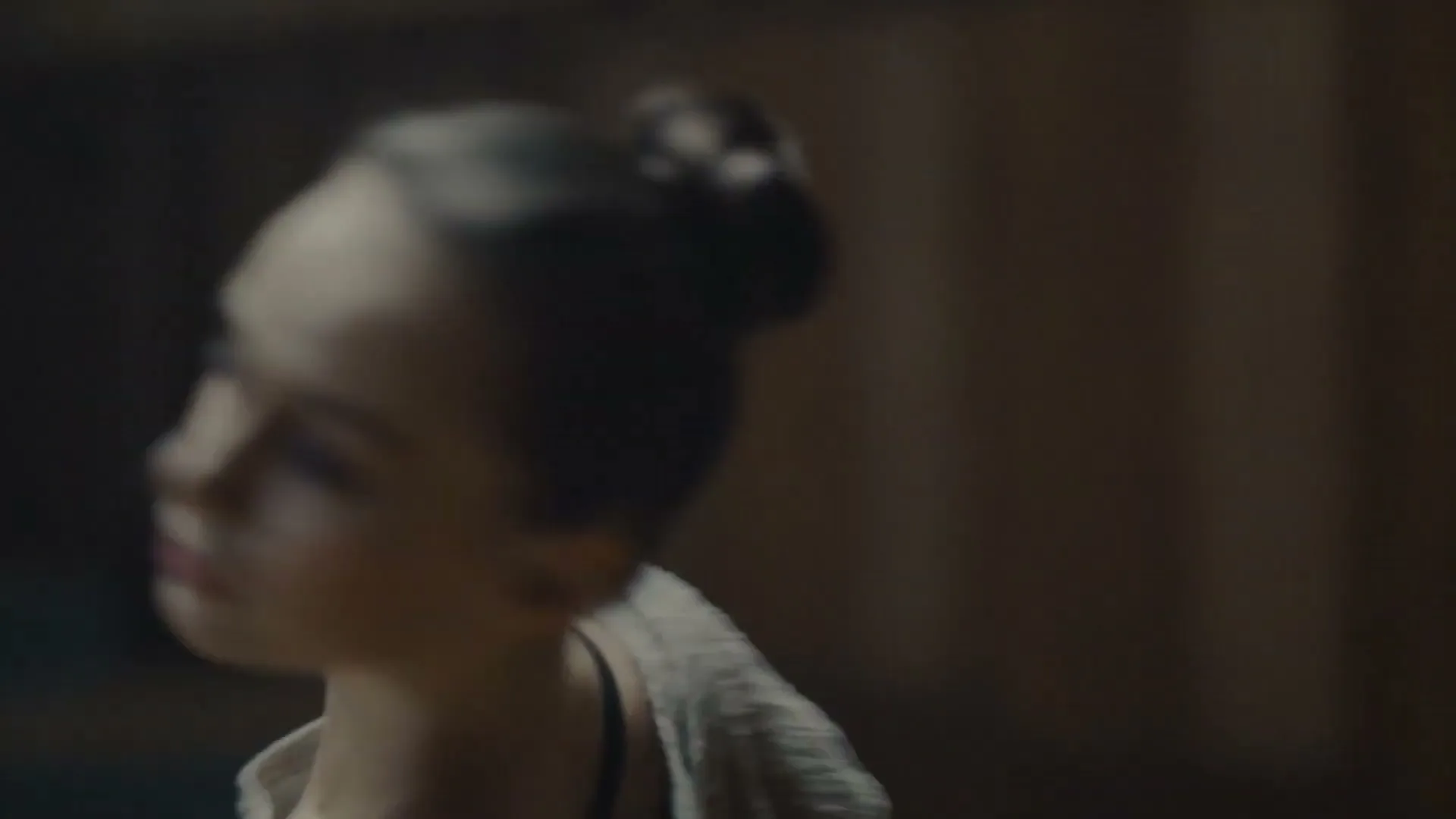 A female ballet dancer with her hair in a bun, wearing a light-colored top, dances gracefully in a dimly lit studio.