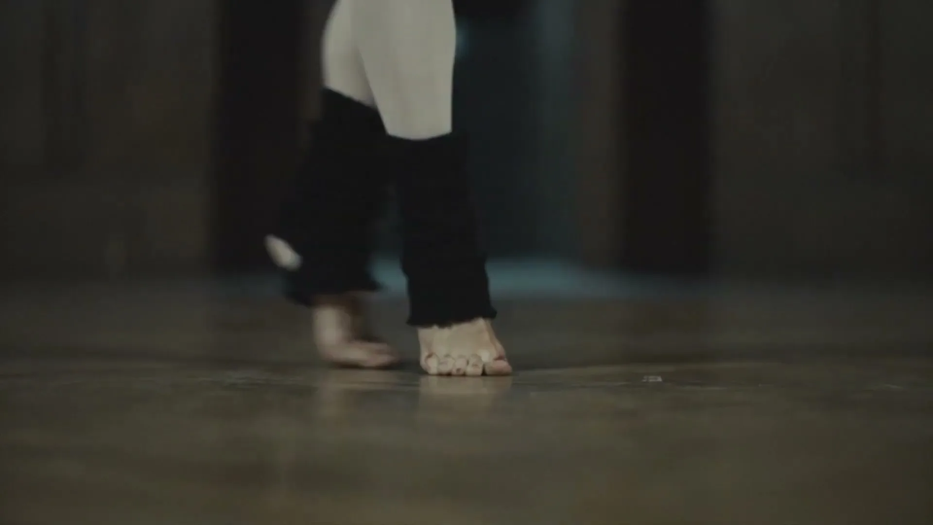 A female ballet dancer's feet, clad in white tights and black leg warmers, are shown on a wooden floor, with a small amount of blood visible on one toe.