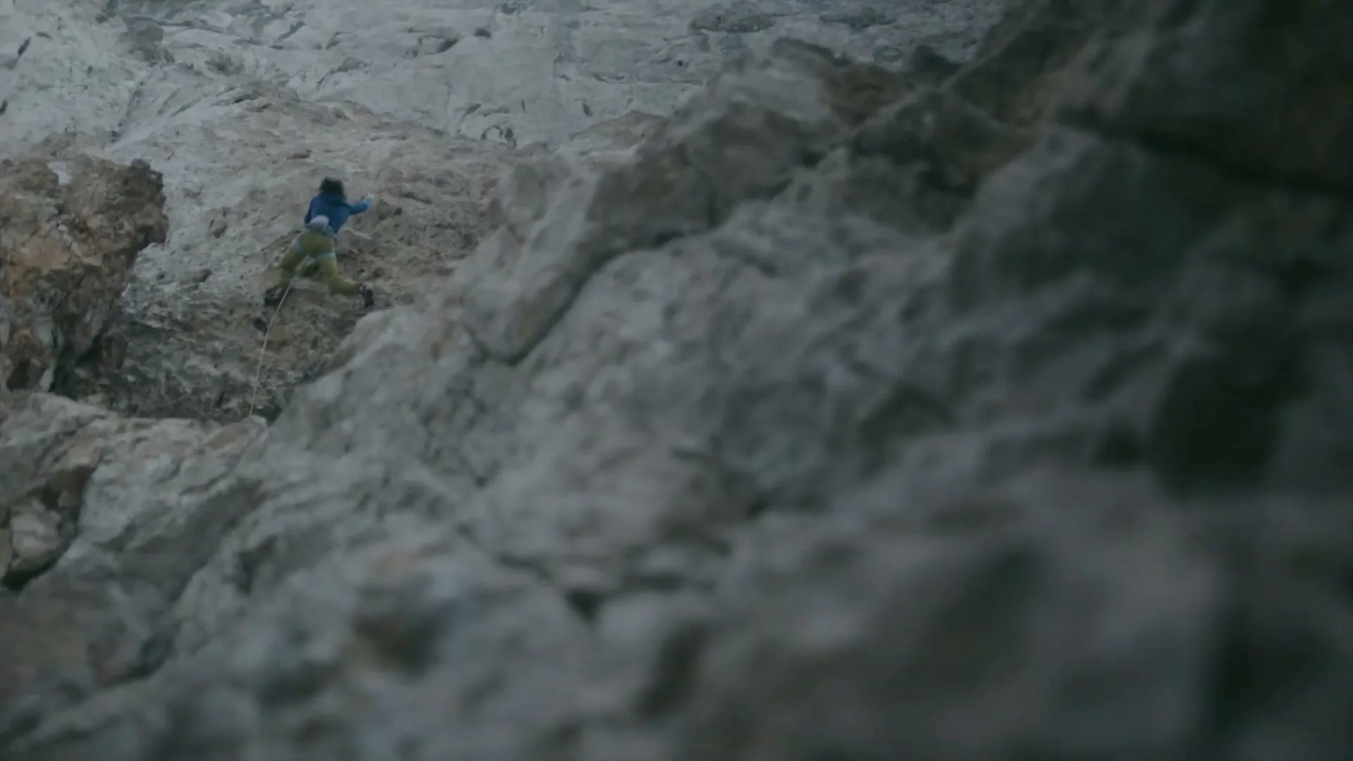 A female rock climber in a blue jacket and green pants scales a steep, rocky cliff face under an overcast sky.