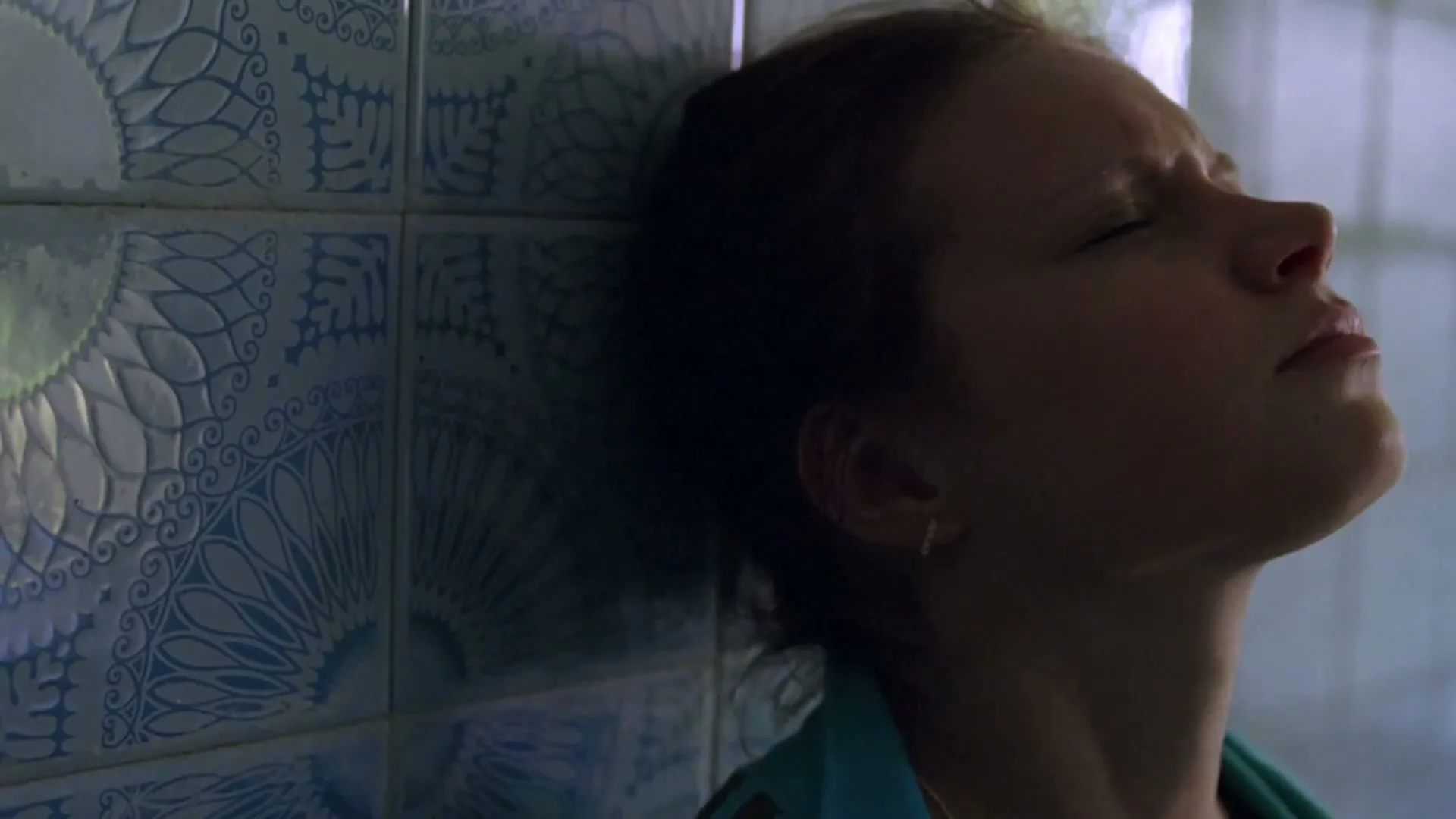 A young woman with dark hair leans against a blue tiled wall, eyes closed, looking pained or contemplative in an interior setting.