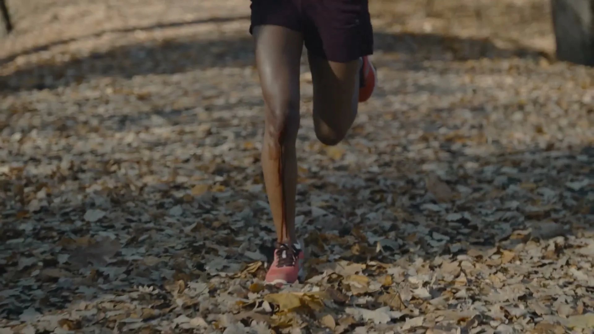 A woman's legs run through a forest path covered in autumn leaves, with a streak of blood visible on her leg.