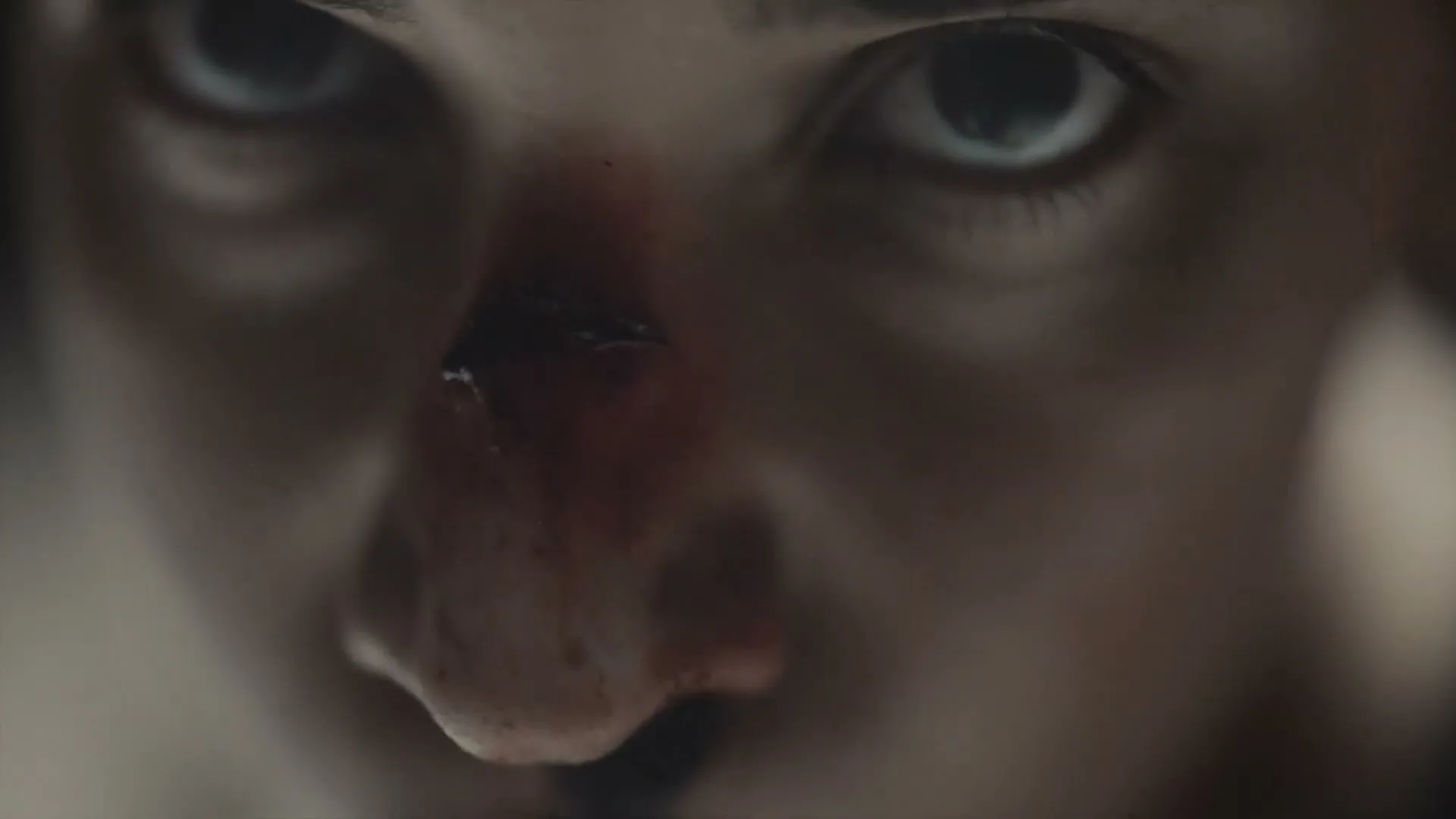 A young woman with a bloody nose looks intensely at the camera, her face filling the frame in a dramatic close-up.