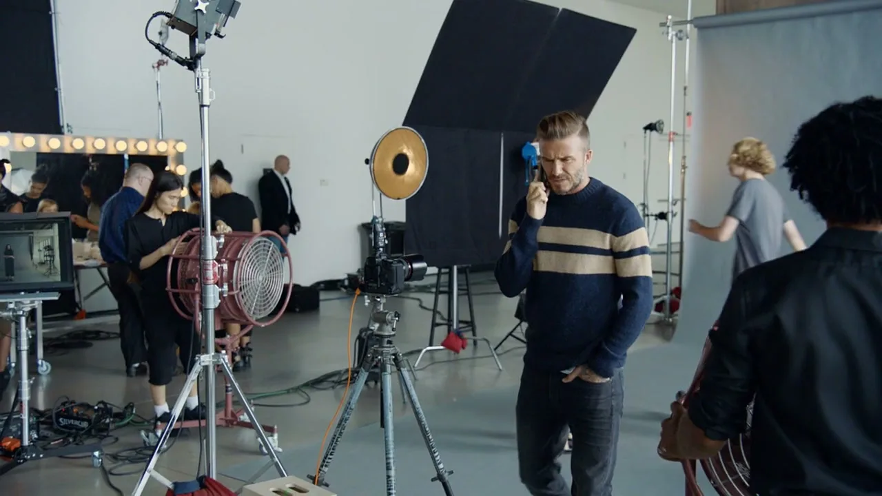 David Beckham, a male in his 40s, wearing a striped sweater and jeans, talks on a phone on a busy film set with crew and equipment.