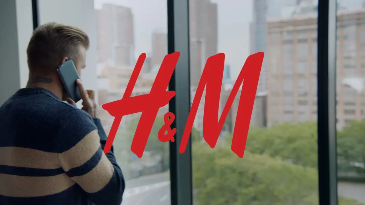 A man with short blonde hair and a striped sweater talks on a phone, looking out a window at a city skyline, with the H&M logo superimposed.