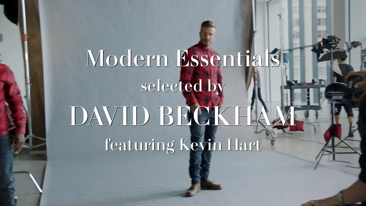 David Beckham, a male in his 40s, wearing a red plaid shirt and jeans, poses for a photoshoot in a bright studio, while Kevin Hart, a male in his 40s, also in a red plaid shirt, walks into the frame.