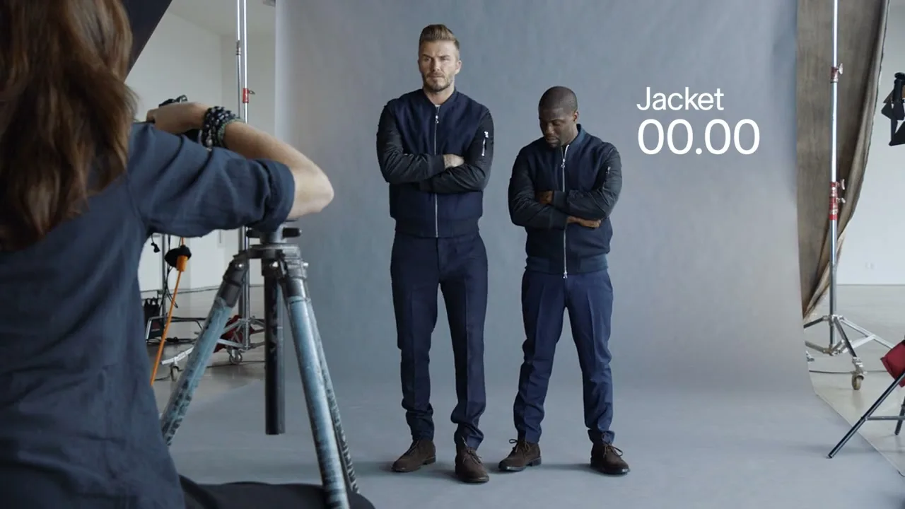 A female photographer shoots David Beckham and Kevin Hart, both wearing matching navy outfits, posing with crossed arms in a bright studio.