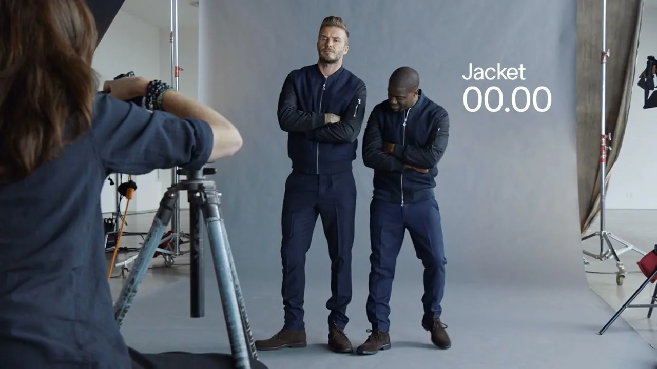 David Beckham and Kevin Hart stand side-by-side in matching outfits, posing for a photoshoot in a studio, while a photographer takes their picture.