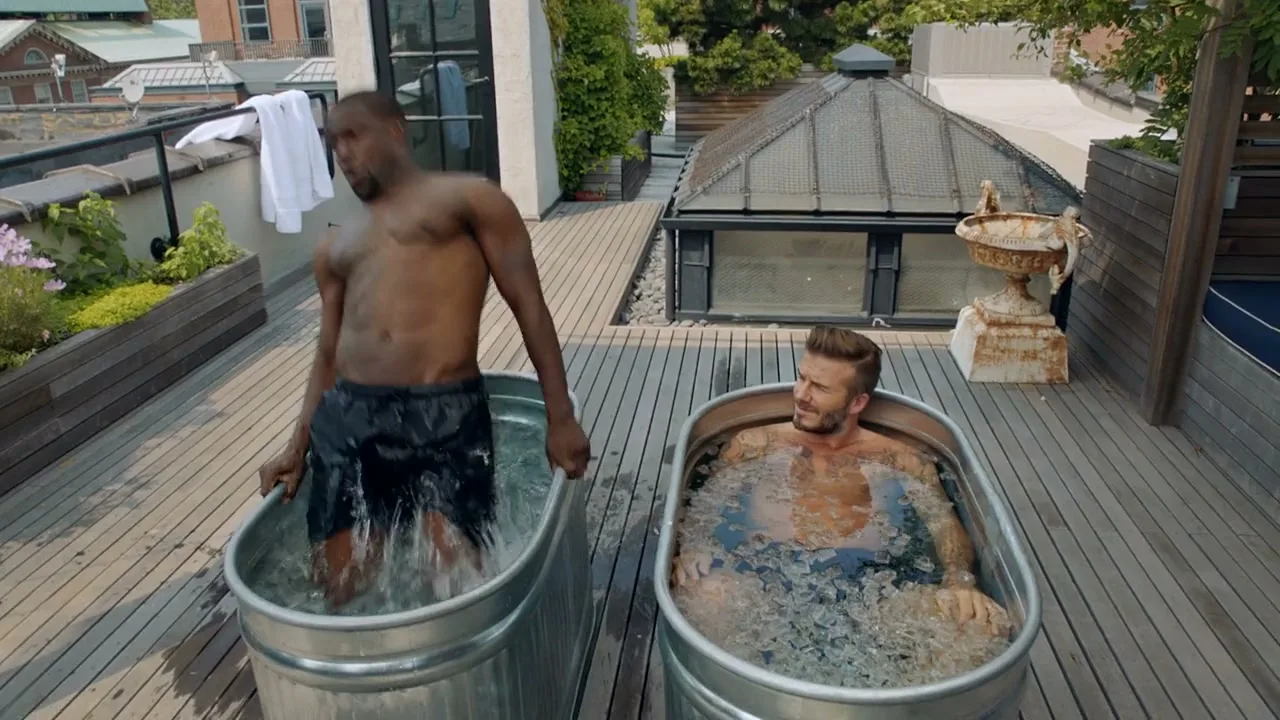 Kevin Hart and David Beckham take an ice bath on a rooftop deck, with Kevin Hart struggling to get into the cold water.