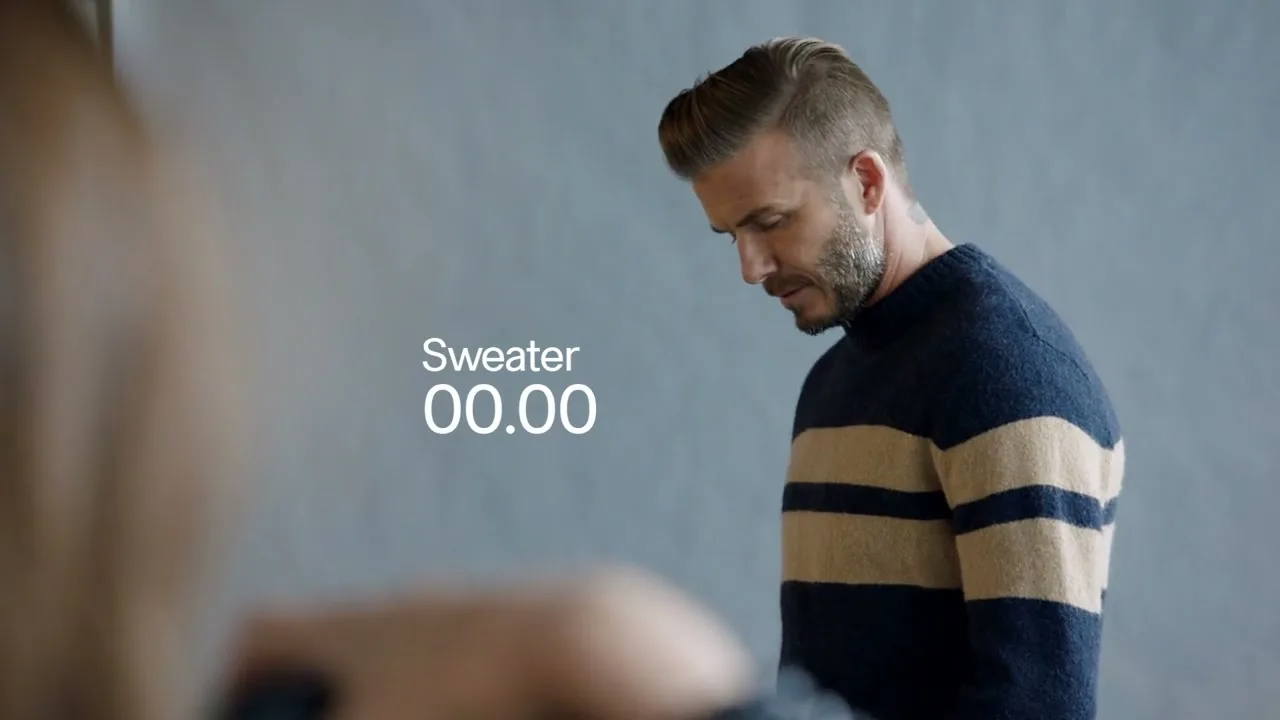 David Beckham, a male in his 40s, wearing a striped sweater, looks down then turns to look at the camera in a studio.