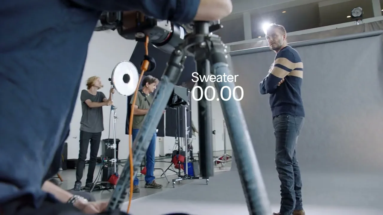 David Beckham, wearing a striped sweater and jeans, stands with arms crossed in a studio while crew members adjust lighting equipment around him.