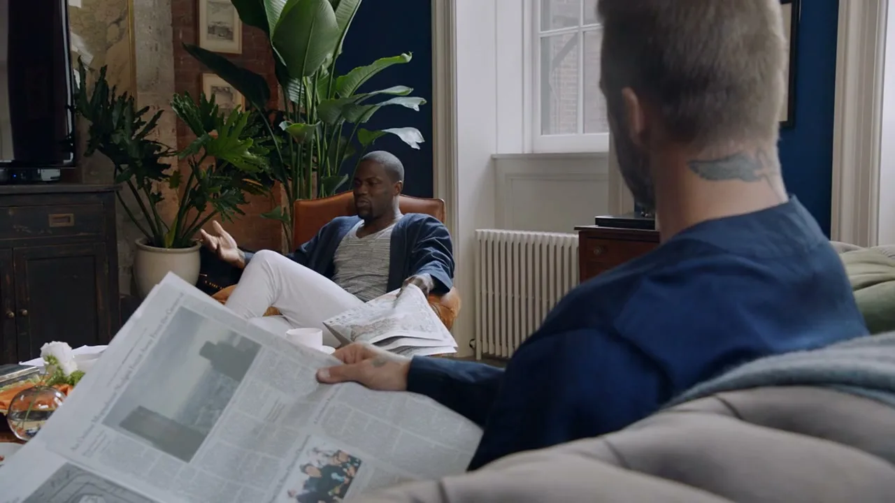 Kevin Hart, dressed in a blue robe and white pants, sits in an armchair, gesturing while talking to David Beckham, who is reading a newspaper in a cozy living room.