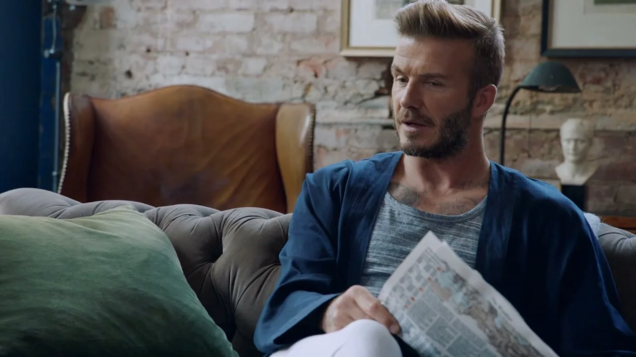 David Beckham, a man with a beard, sits on a couch holding a newspaper in a rustic living room.