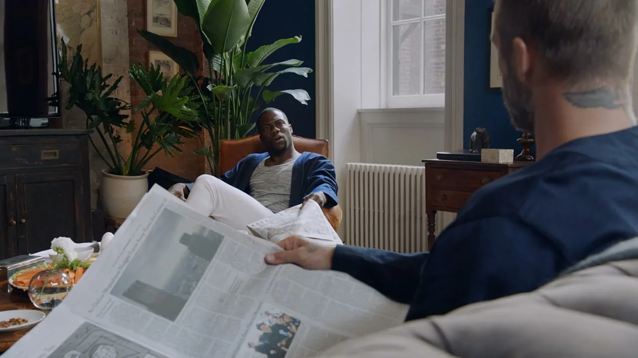 Kevin Hart, dressed in a blue robe and white pants, sits in a brown leather chair, looking at a man reading a newspaper in a well-lit living room.