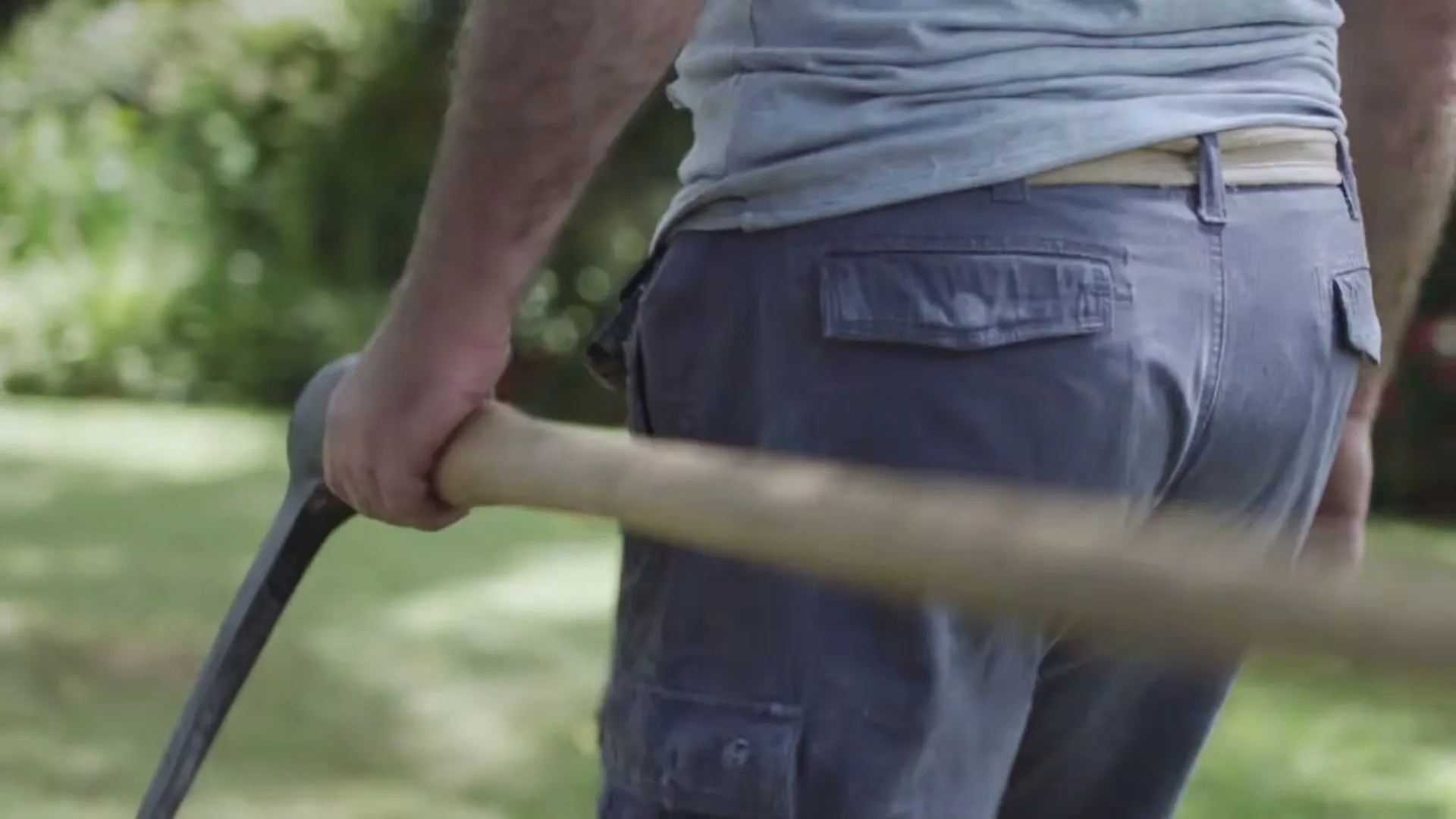 A man in blue shorts and a grey t-shirt walks through a sunny garden, carrying a pickaxe over his shoulder.