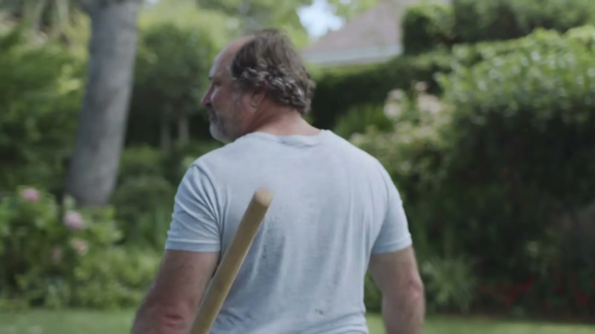 A middle-aged man with a receding hairline and a light blue t-shirt walks away from the camera, carrying a wooden stick over his shoulder in a lush green garden.