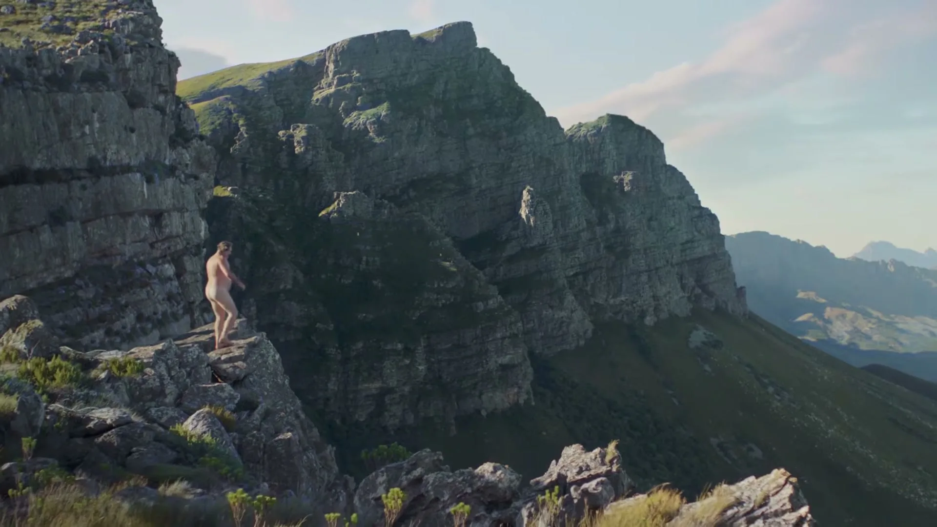 A naked man walks along a rocky mountain ridge, looking out at the vast landscape under a bright sky.