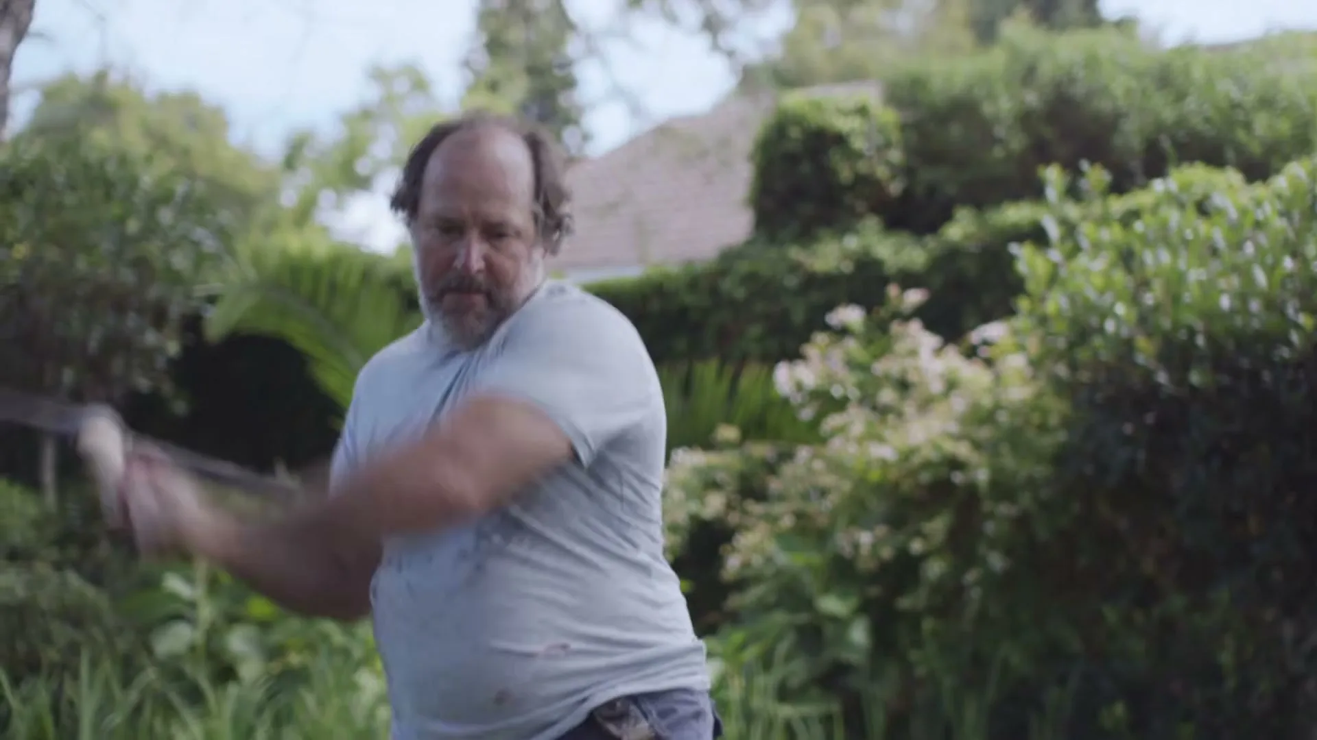 A disheveled man with a beard and dirty t-shirt swings a pickaxe in a lush green garden under a bright sky.