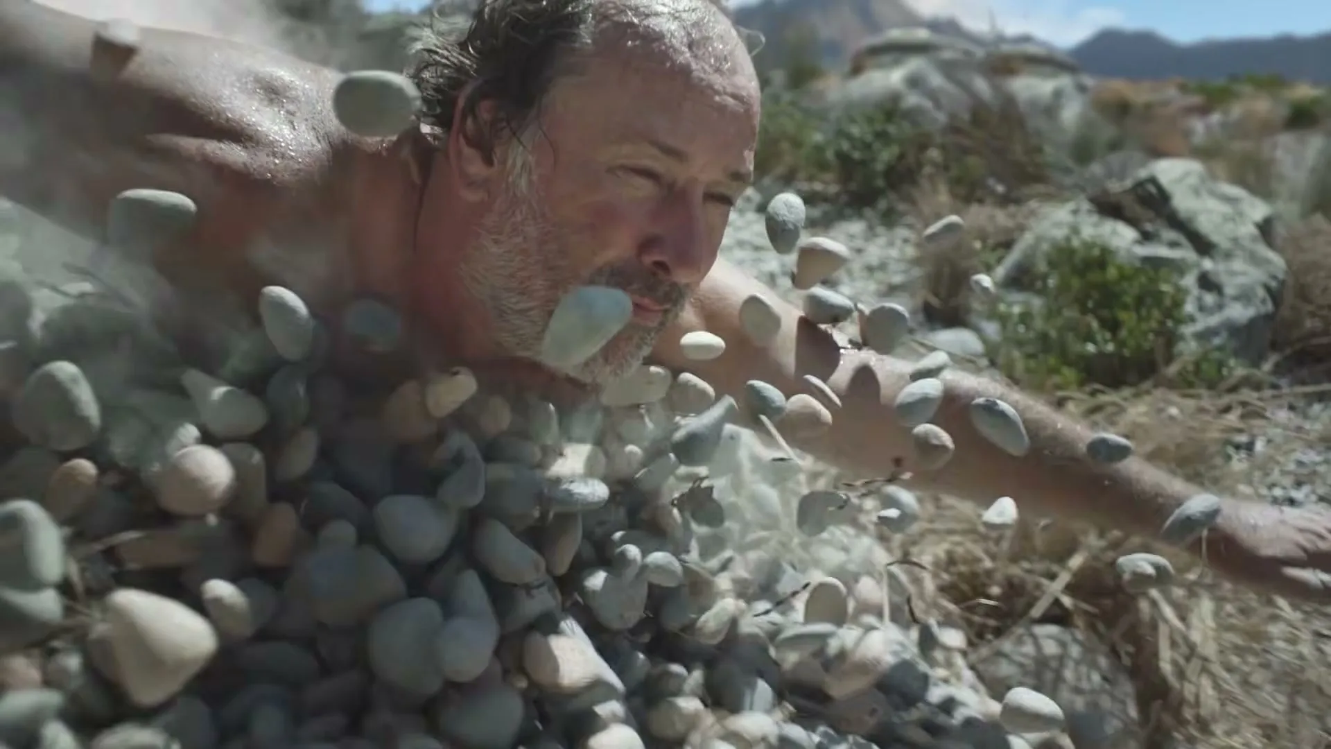 A shirtless man with a beard is buried in small rocks, with only his head and arms visible, as rocks fly around him in a dry, rocky landscape.