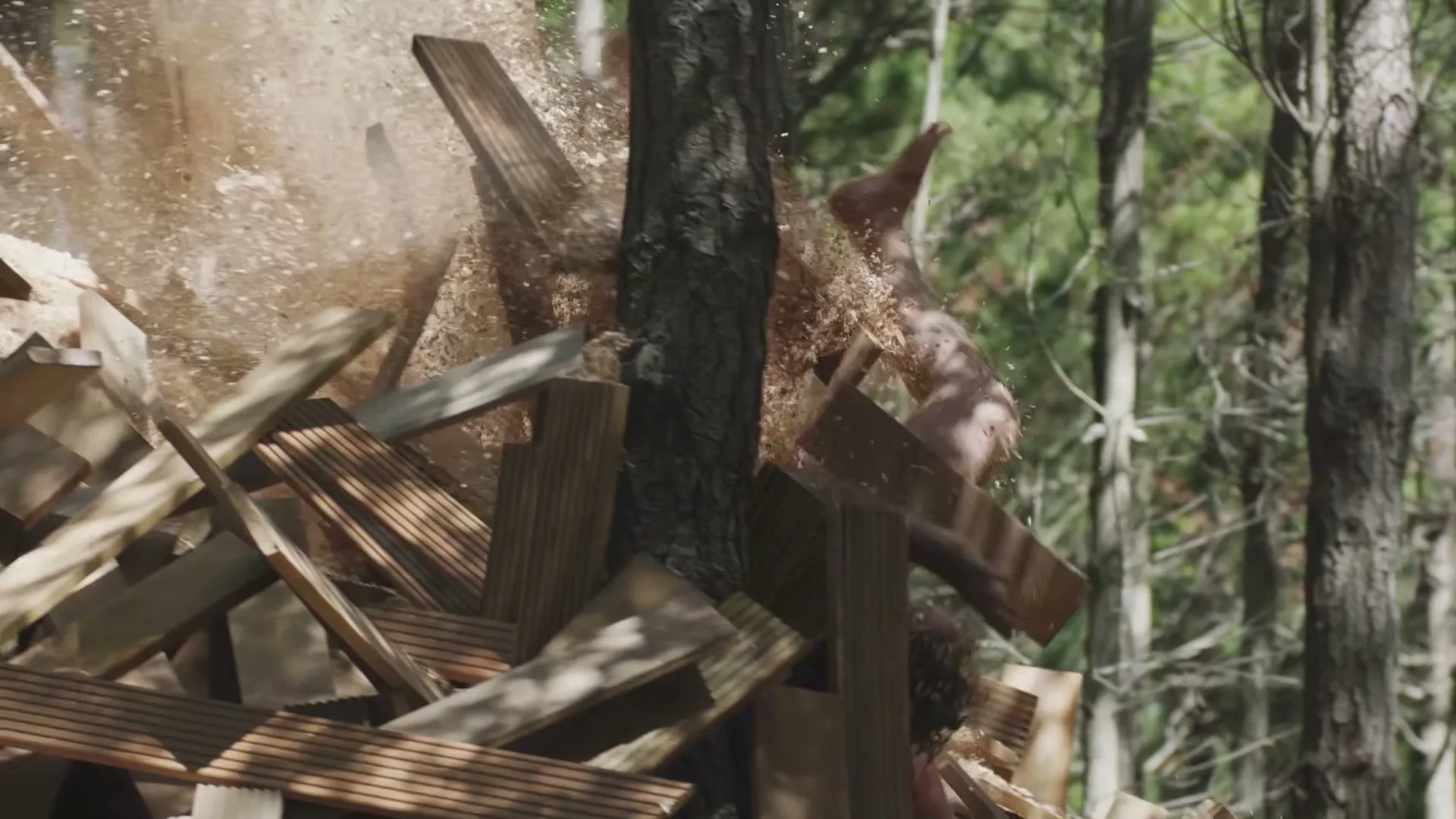 A person falls into a pile of wood planks and sawdust in a forest, with trees visible in the background.