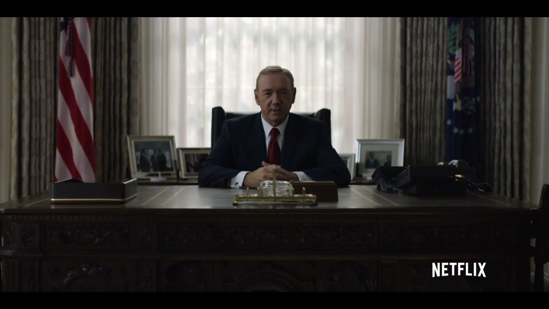 A male politician in a suit sits at a large wooden desk in an office, looking directly at the camera with a serious expression.