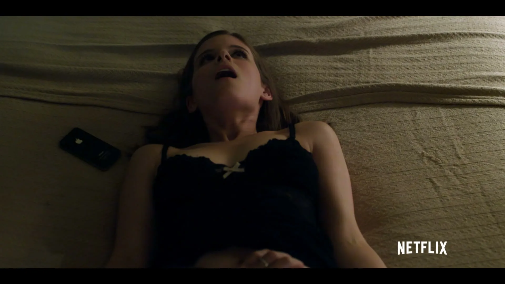 A woman in a black nightgown lies on a bed, looking up with her mouth open, with a phone next to her.