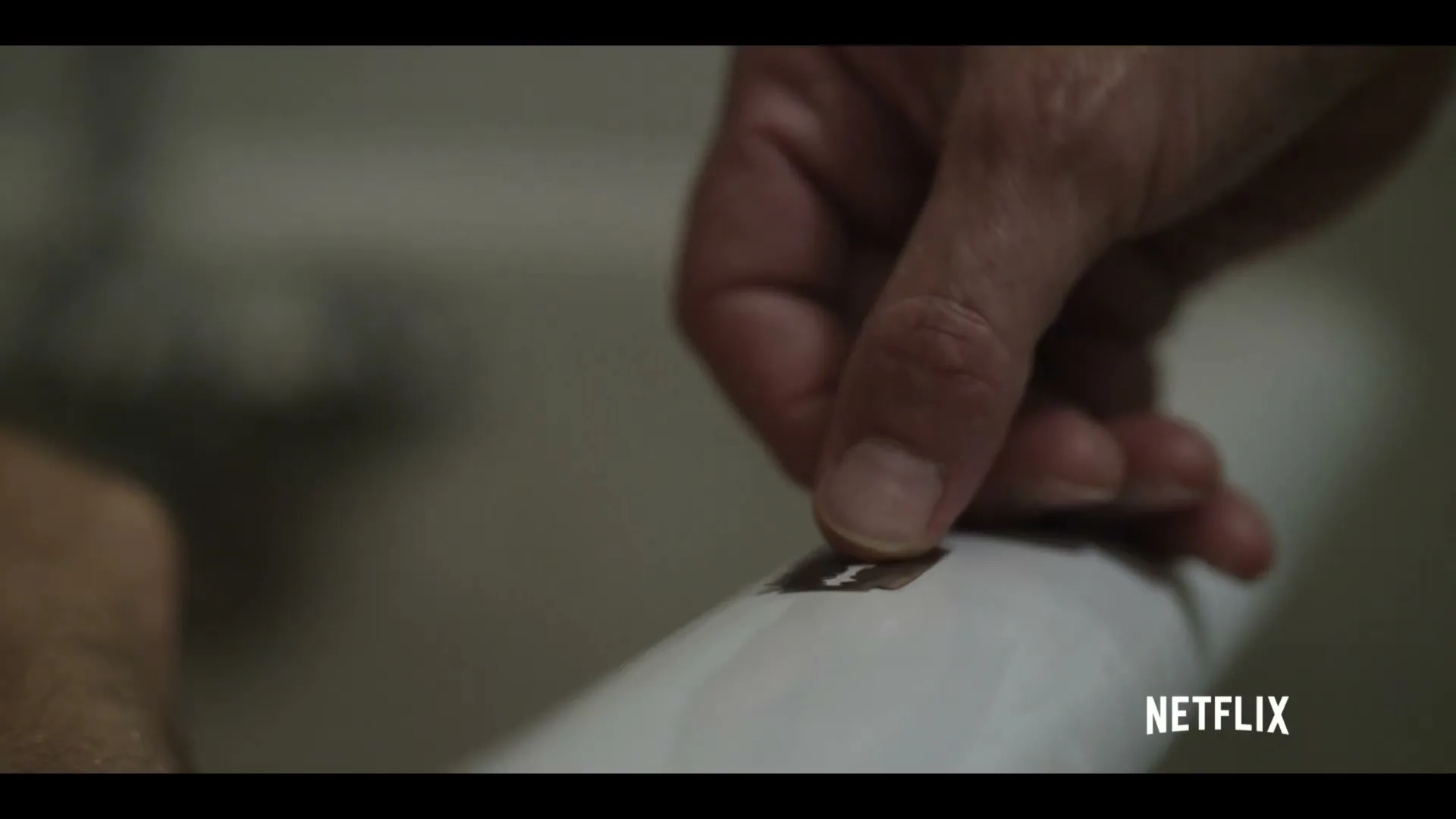 A man's hand carefully places a razor blade onto a white surface, possibly a toilet or sink, in a dimly lit bathroom.