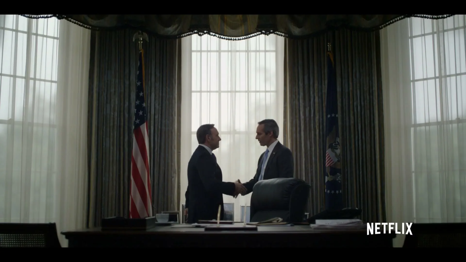 Two men in suits shake hands in a formal office with large windows and flags, suggesting a political setting.