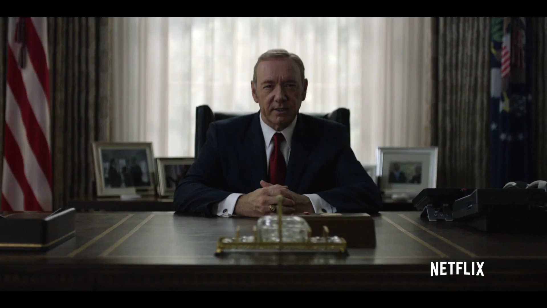 A man in a suit sits behind a large desk, looking directly at the camera in a formal office setting.