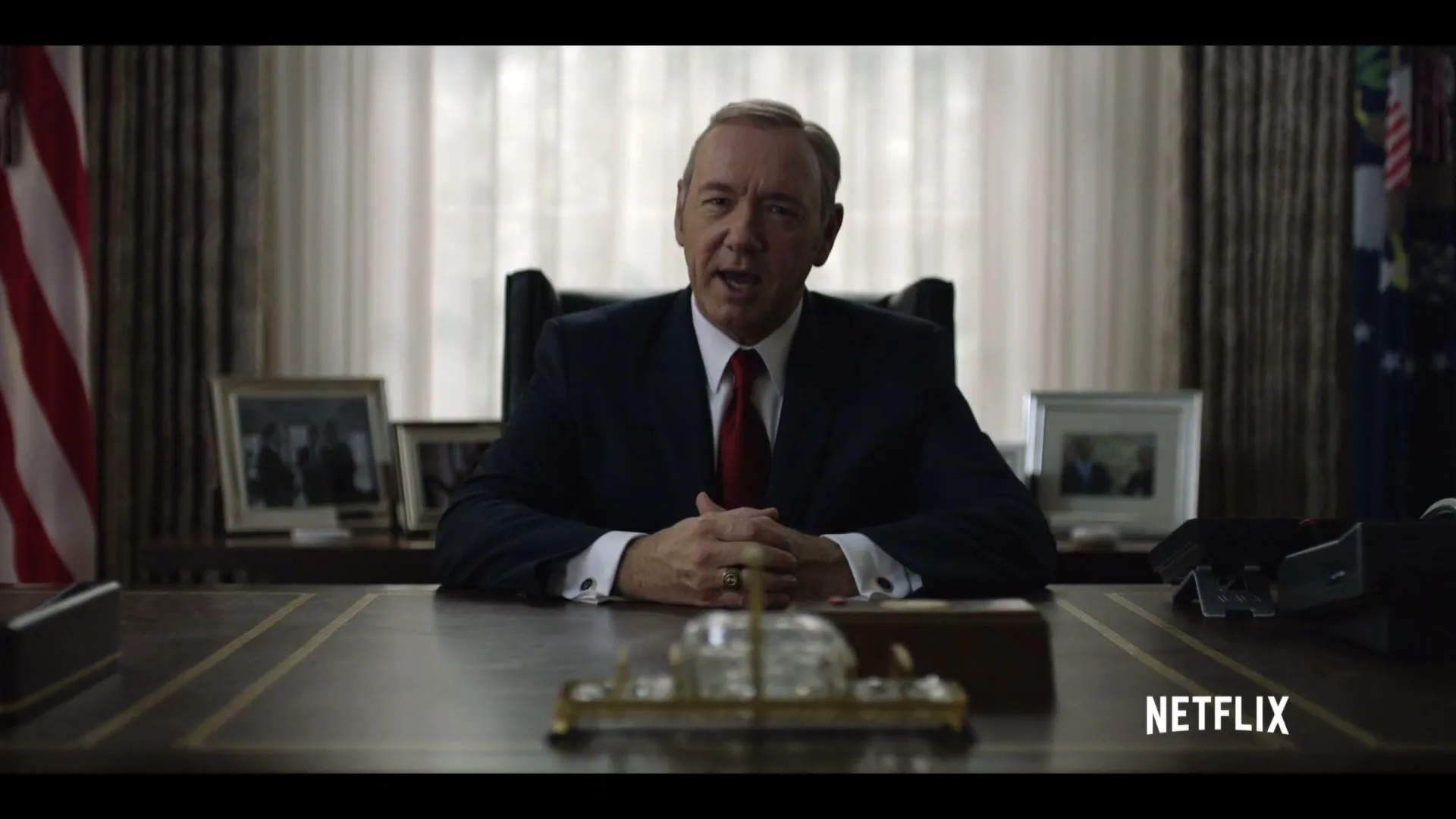 A man in a suit sits behind a large desk, looking directly at the camera, with flags and framed photos in the background.