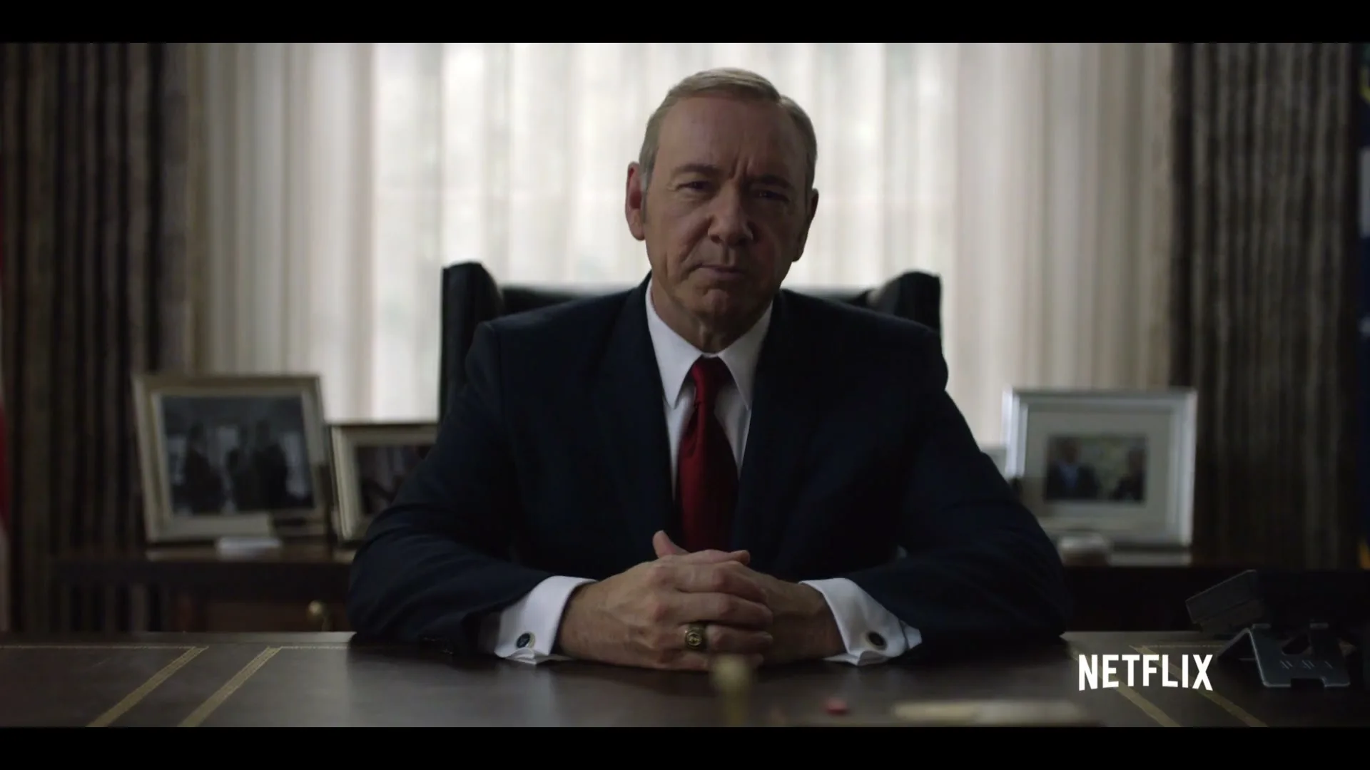 A man in a suit sits at a large desk, looking directly at the camera, in an office setting.