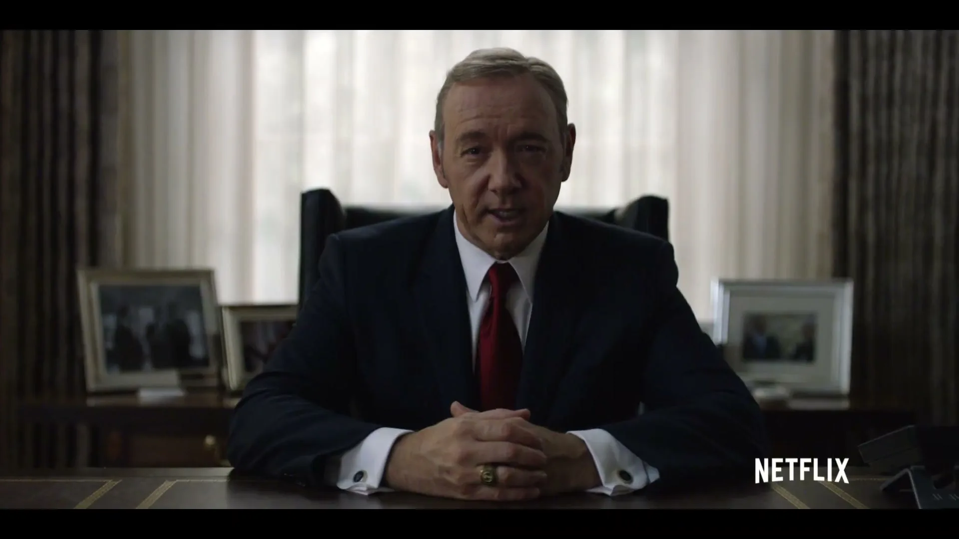 A man in a suit sits at a desk, looking directly at the camera in an office.