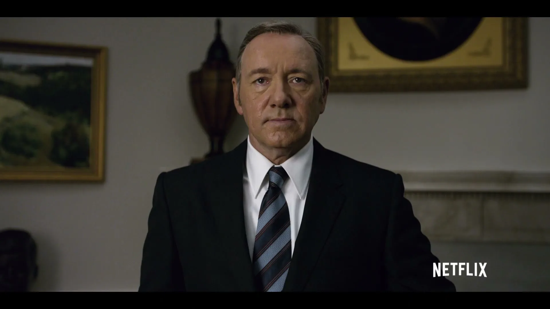A middle-aged man in a suit and tie looks directly at the camera in a formal room with paintings on the wall.