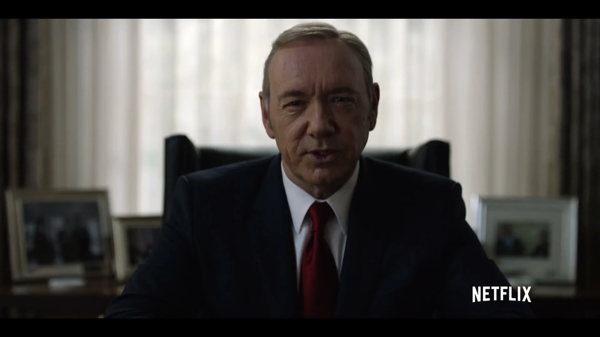 A man in a suit and red tie sits at a desk, looking directly at the camera and speaking in a serious tone in an office.