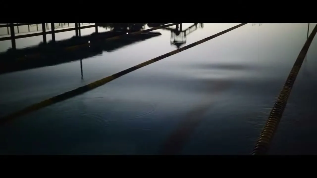 A wide shot of a dark, still swimming pool with lane lines, as ripples appear on the water's surface, reflecting the surrounding structures.