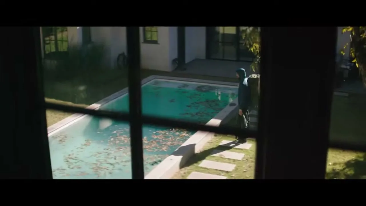 A man in a dark tracksuit stands by a neglected swimming pool in a suburban backyard, viewed through a window.