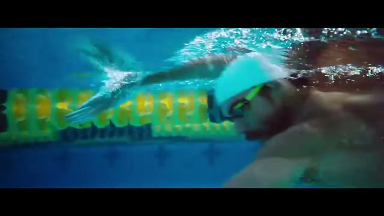 An underwater shot shows a male swimmer in a white cap and green goggles swimming freestyle next to a yellow lane divider in a pool.