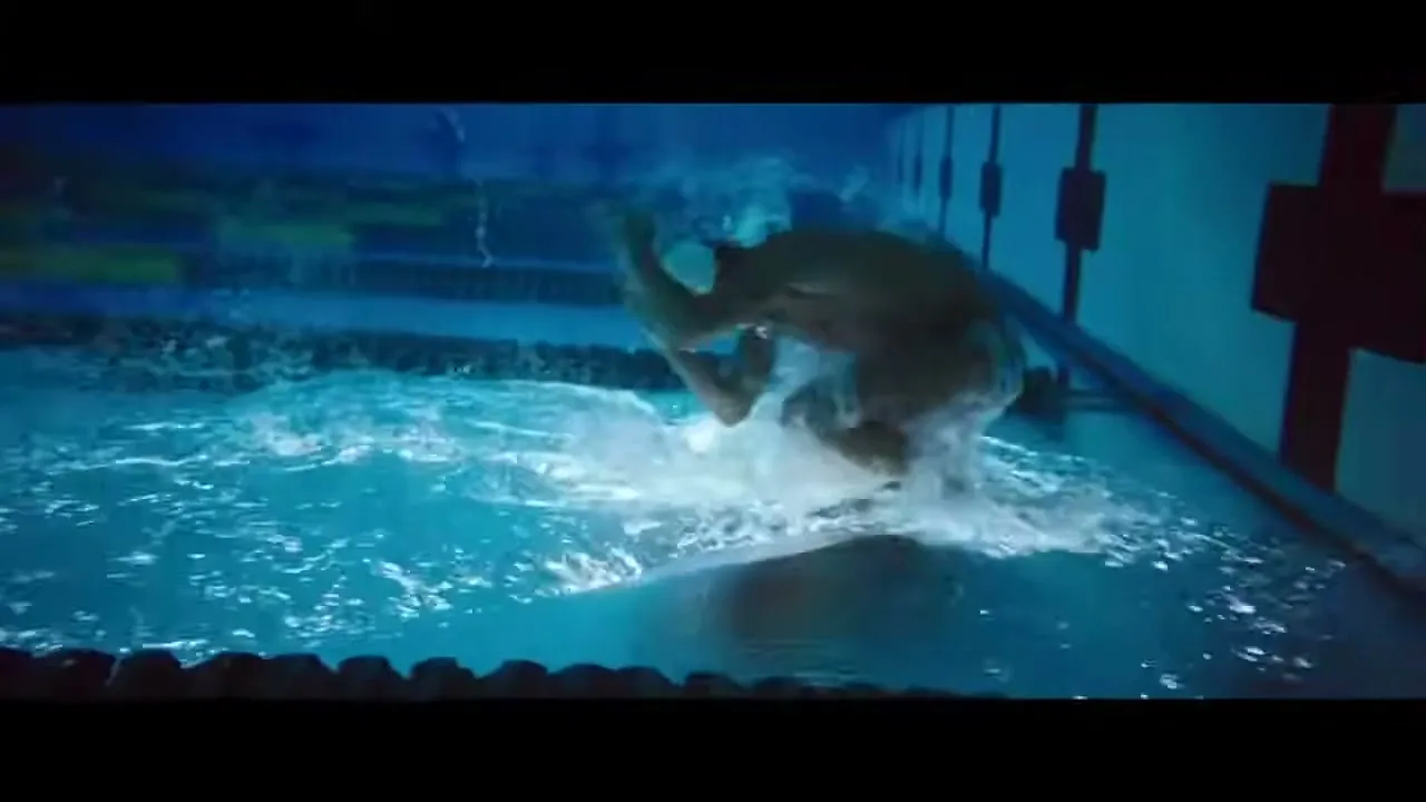 A male swimmer in a cap and briefs pushes off the wall underwater, creating splashes in a dark indoor pool.