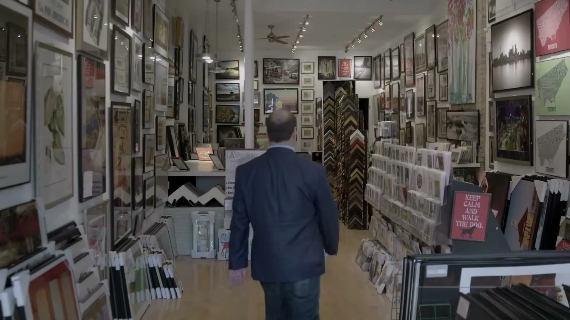A bald man in his 40s, wearing a blue blazer and jeans, walks away from the camera down a narrow aisle in a brightly lit art gallery.