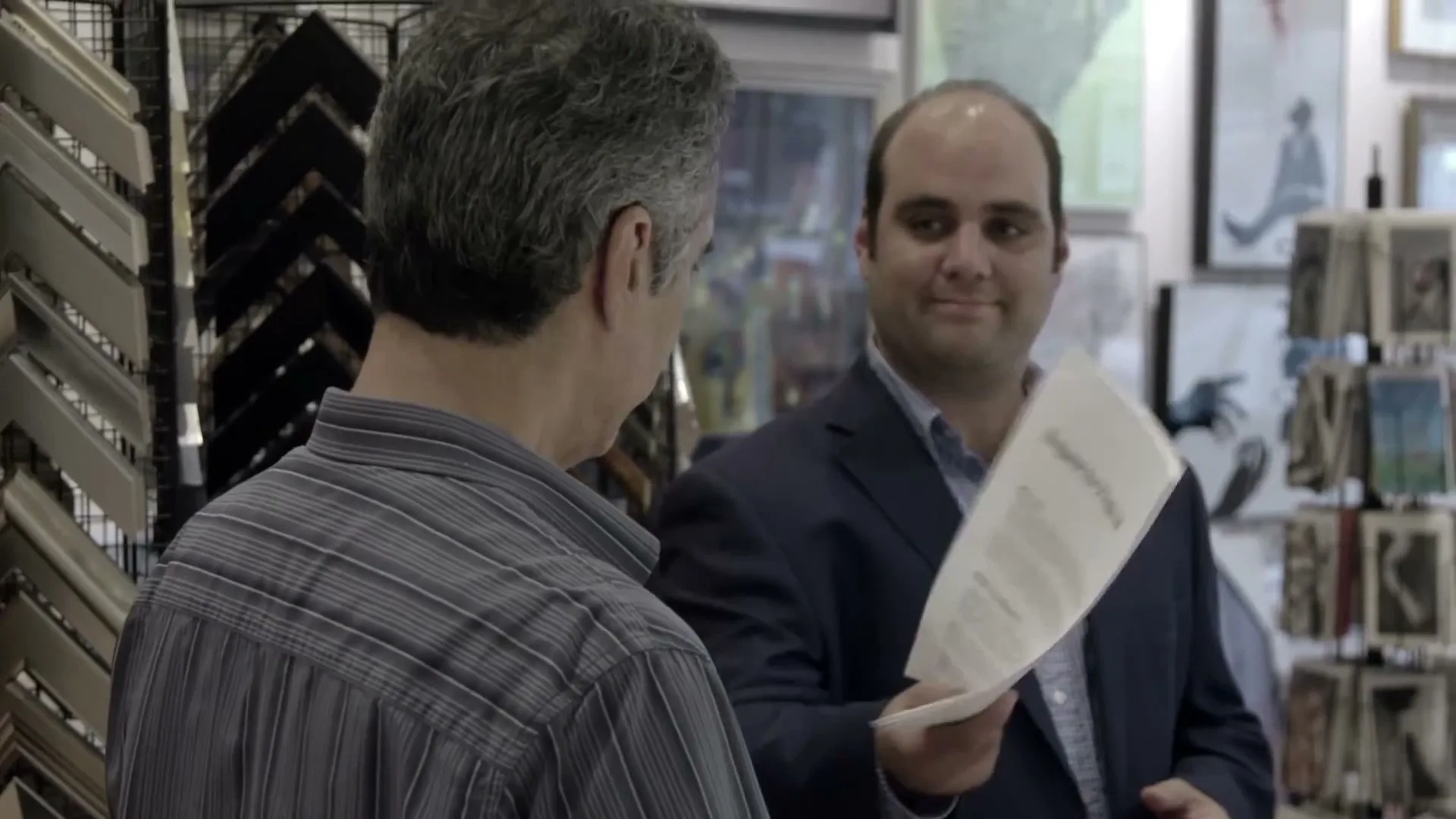 A man in a blue suit holds out a "Request for Proposal" document to another man in a striped shirt, who looks at it with a confused expression in a frame shop.