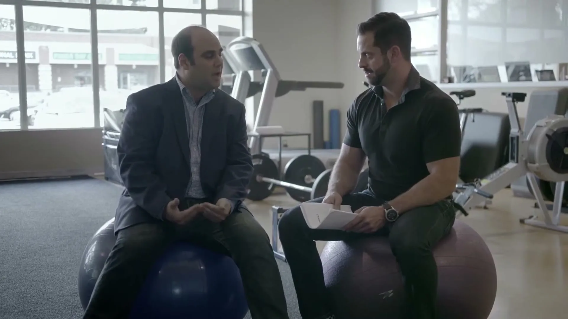 Two men, one in a suit and one in athletic wear, sit on exercise balls in a gym, talking to each other.