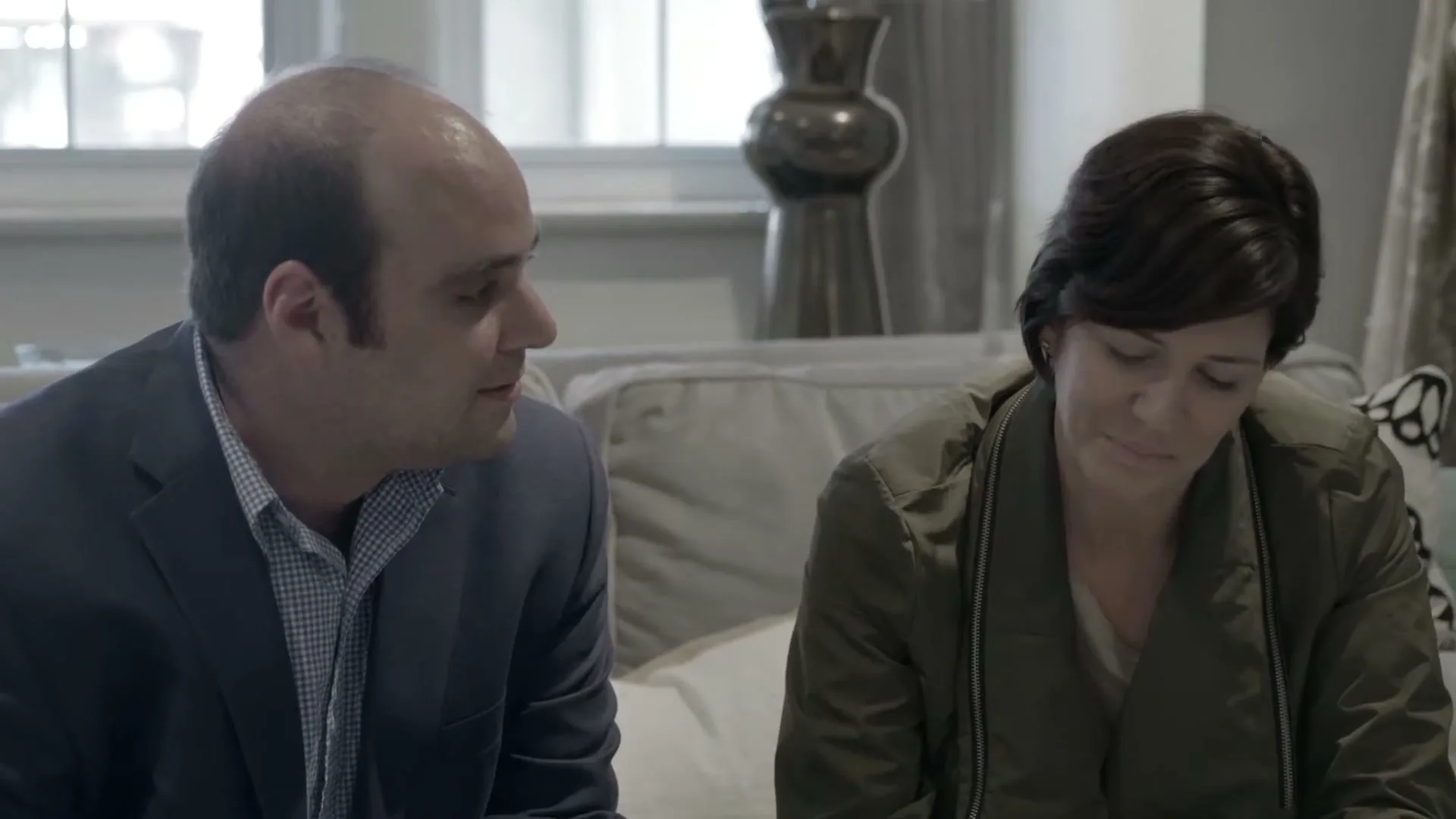 A bald man in a suit talks to a woman with short hair in a jacket, who looks down then up at him, in a living room.