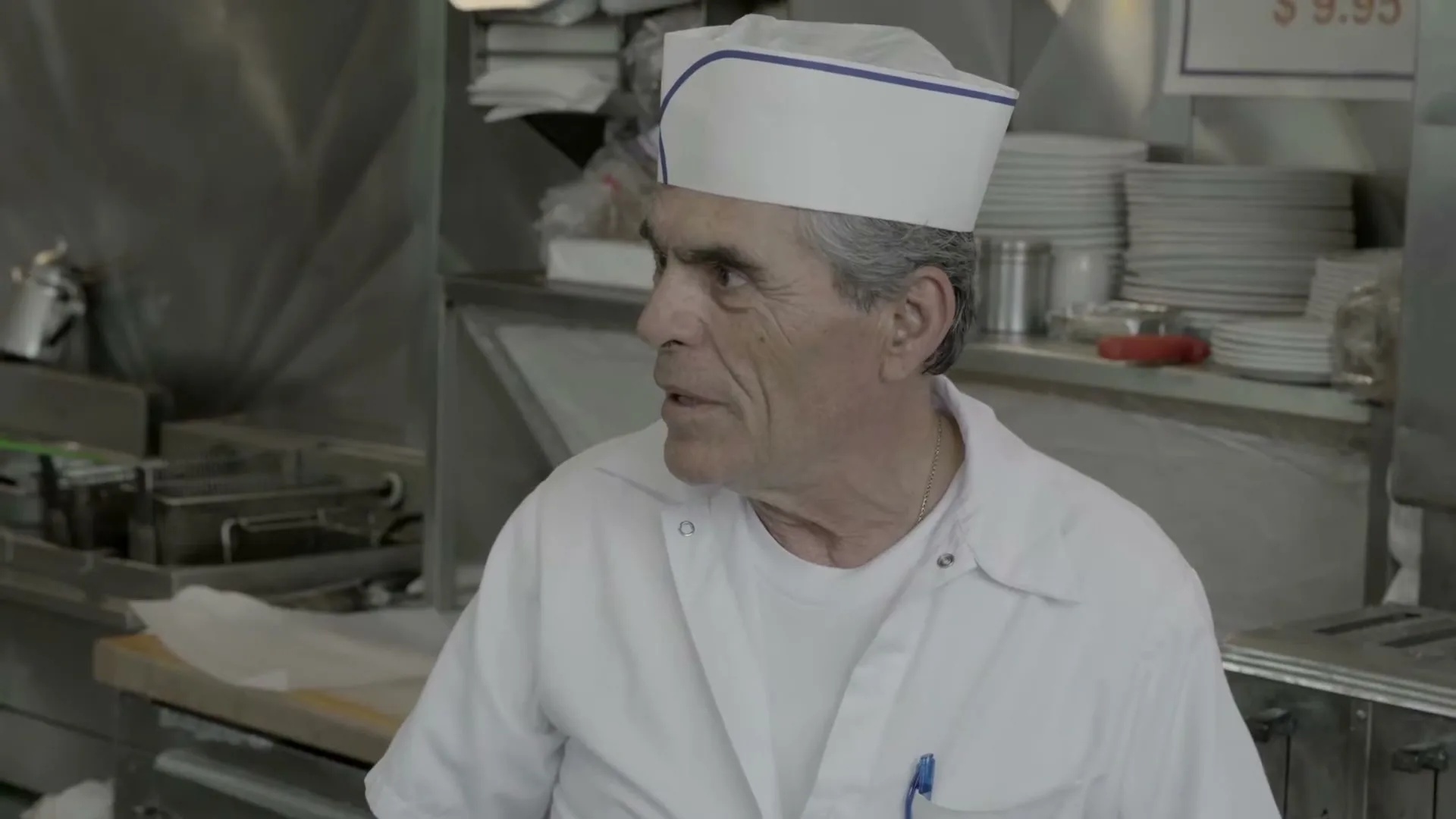 A male chef in his 60s, wearing a white uniform and paper hat, looks off-camera with a surprised expression in a busy commercial kitchen.