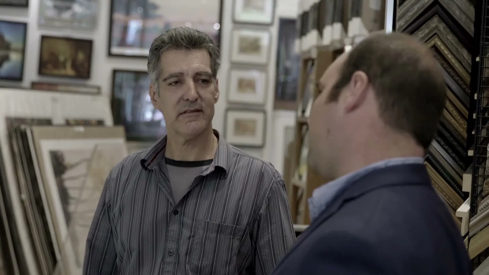 A middle-aged man with grey hair and a striped shirt talks to another man in a frame shop, surrounded by various framed art and empty frames.