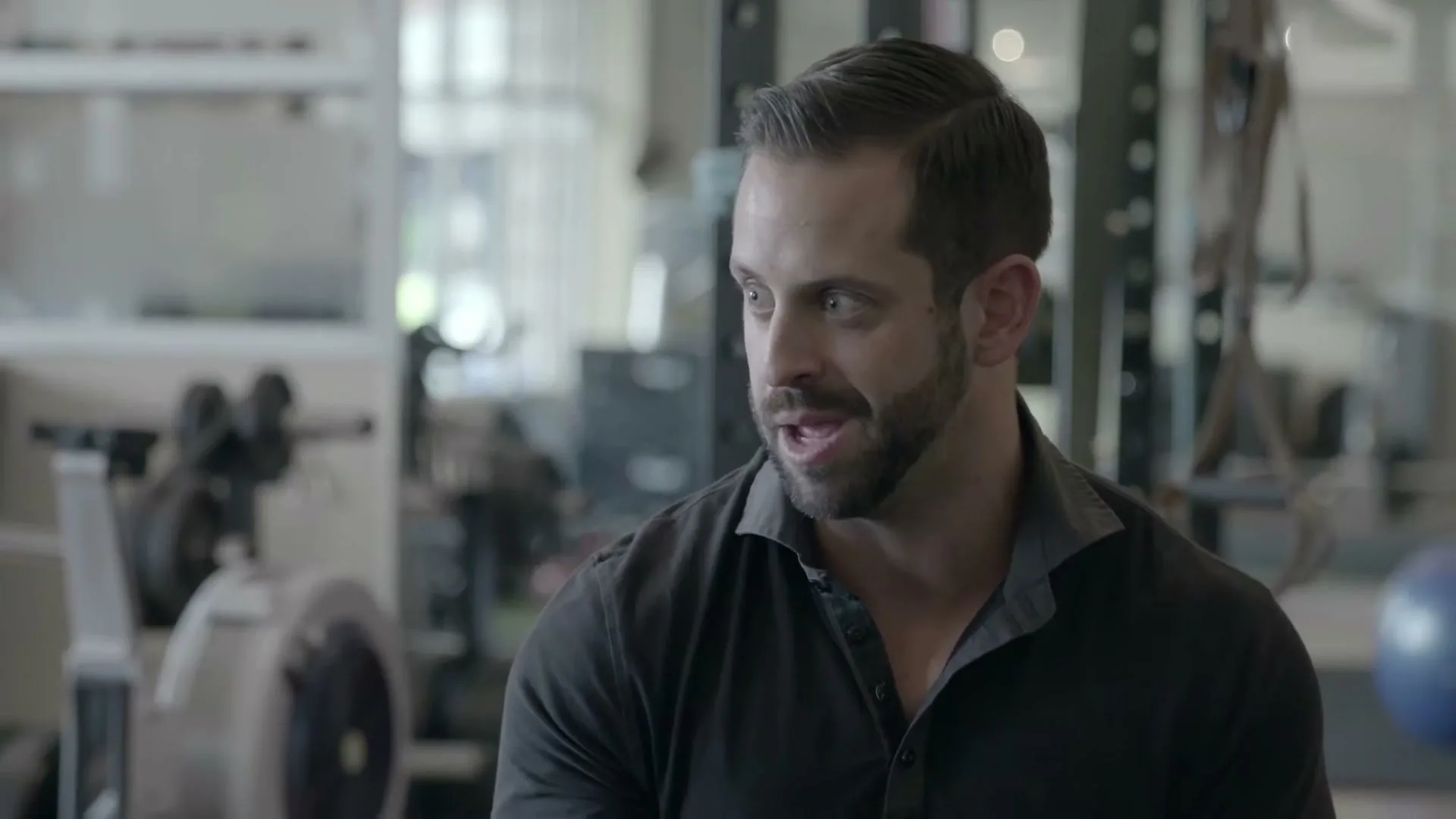 A fit man with a beard and dark shirt looks around and speaks in a gym, with exercise equipment visible in the background.