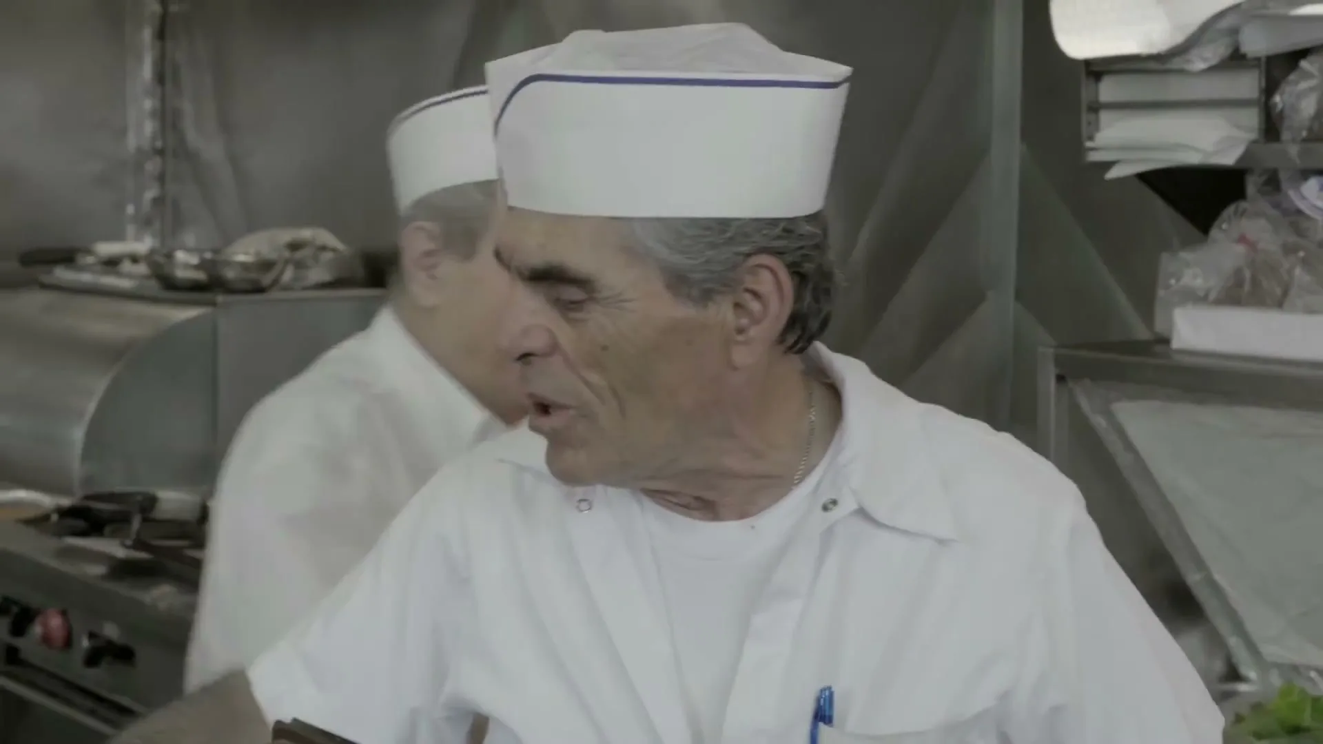 A male chef in his 60s, wearing a white uniform and paper hat, talks animatedly in a busy commercial kitchen.