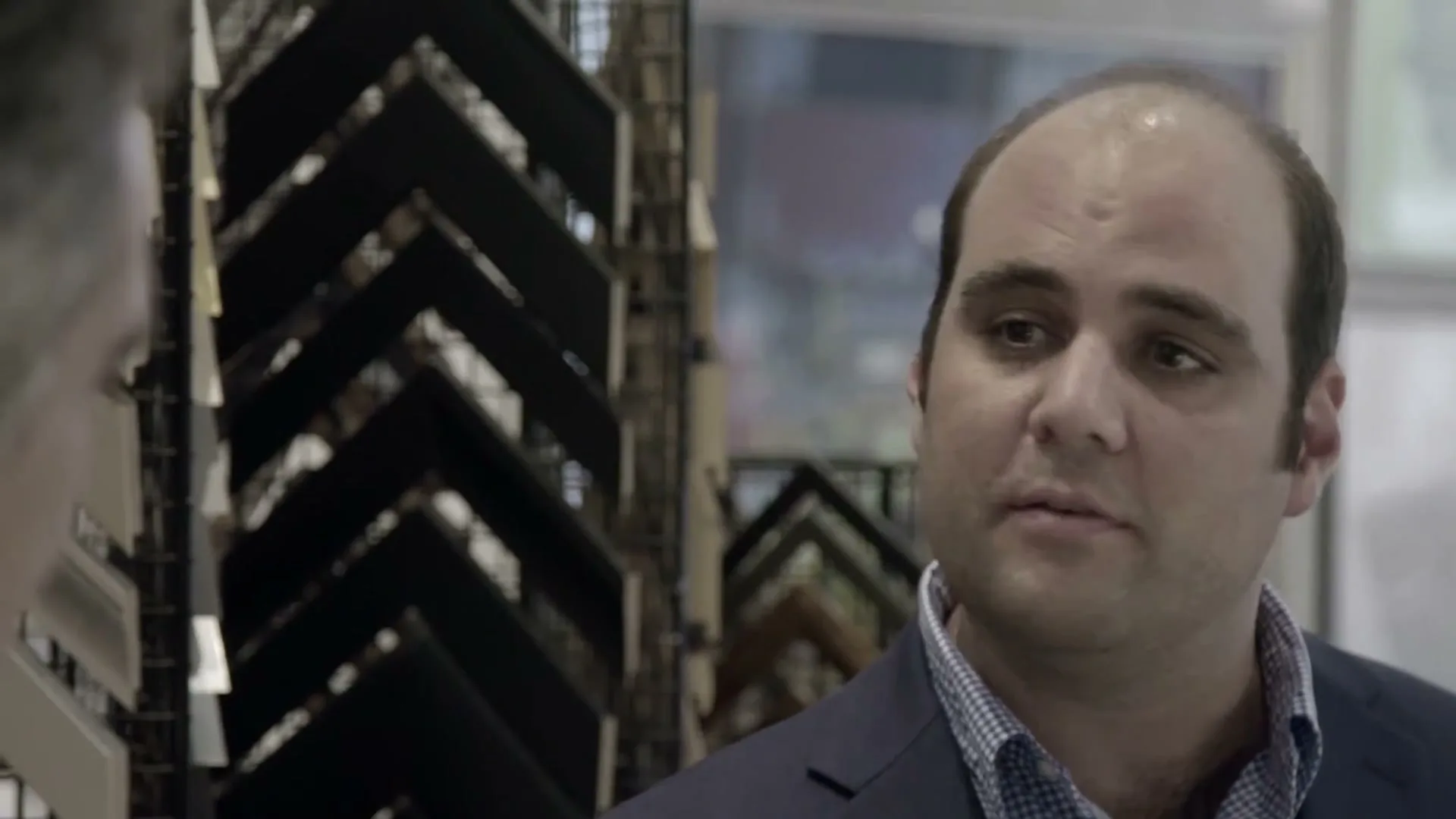 A balding man in his 30s, wearing a blue suit jacket and checkered shirt, looks at another person off-screen in a store.