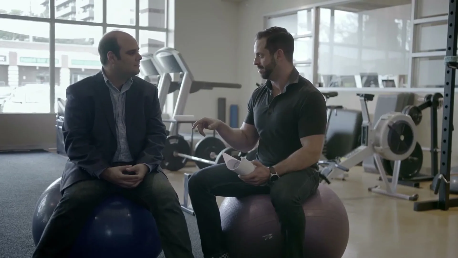 Two men sit on exercise balls in a gym, talking to each other, with one gesturing while holding papers.
