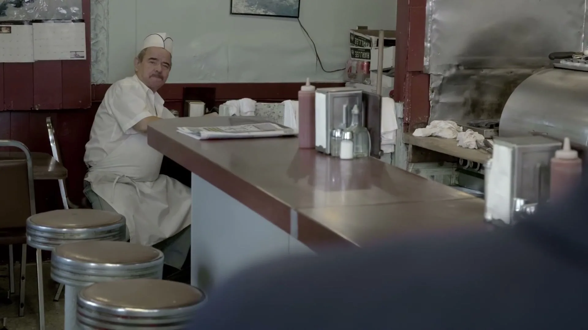 A male chef in his 50s, wearing a white uniform, sits at a diner counter, looking at a newspaper, then looks up at someone off-screen.