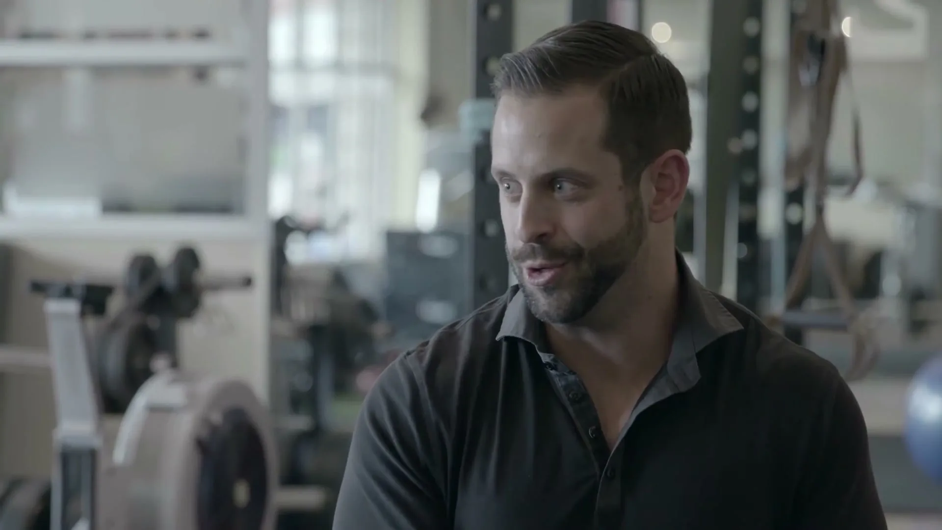 A man with a beard and dark shirt looks to the side and talks in a gym setting.